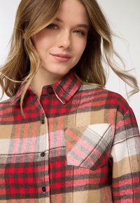 Camicia di flanella a quadri rossi, beige e marroni con chiusura frontale con bottoni, colletto e taschino sul petto caratterizzato da un motivo diagonale.