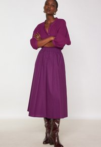 Maglietta lunga manica a coste viola con bottoni, abbinata a una gonna maxi con vita elastica dello stesso colore e stivali marroni al ginocchio.