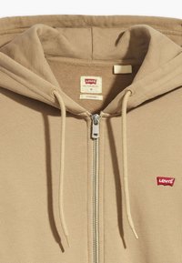 Beige dragkedja hoodie gjord av mjukt tyg, med fodrad huva, snören och en liten röd Levi's-logotyp på bröstet.