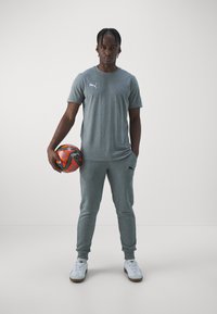 Grå treningst-skjorte og matchende joggebukser med elastiske mansjetter, kombinert med hvite joggesko. Personen holder en oransje og svart fotball.