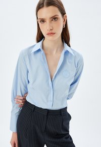 Camisa de manga larga azul claro con cuello puntiagudo, botón frontal y detalle bordado en el pecho, combinada con pantalones oscuros de rayas.