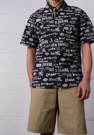 Person trägt ein schwarzes kurzärmeliges Hemd mit weißen "Dickies"-Texten über das gesamte Hemd und beige knielange Shorts.