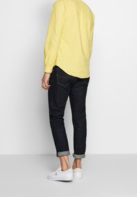 Żółta koszula z długim rękawem, ciemne jeansy z podwiniętym mankietem oraz białe trampki, widziane od tyłu na neutralnym tle.