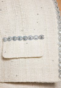 Țesătură albă din tweed cu un țesătură texturată, având un tiv decorativ argintiu și broderie pe un buzunar frontal.