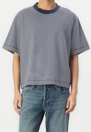 Person trägt ein navy-blaues und weiß gestreiftes T-Shirt mit kurzen Ärmeln und blaue Jeans und steht vor einem schlichten hellen Hintergrund.