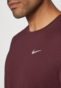 Burgunderfarget Nike-skjorte med rund hals, laget av lett stoff. Har en liten sølvfarget Nike-swoosh-logo på venstre bryst.