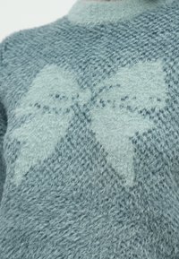 Ανοιχτό μπλε πουλόβερ κατασκευασμένο από απαλό, αφράτο υλικό, με μεγάλο μοτίβο φιόγκου σε ελαφρώς πιο σκούρο μπλε. Στρογγυλή λαιμόκοψη και υφή με ανάγλυφη επιφάνεια.