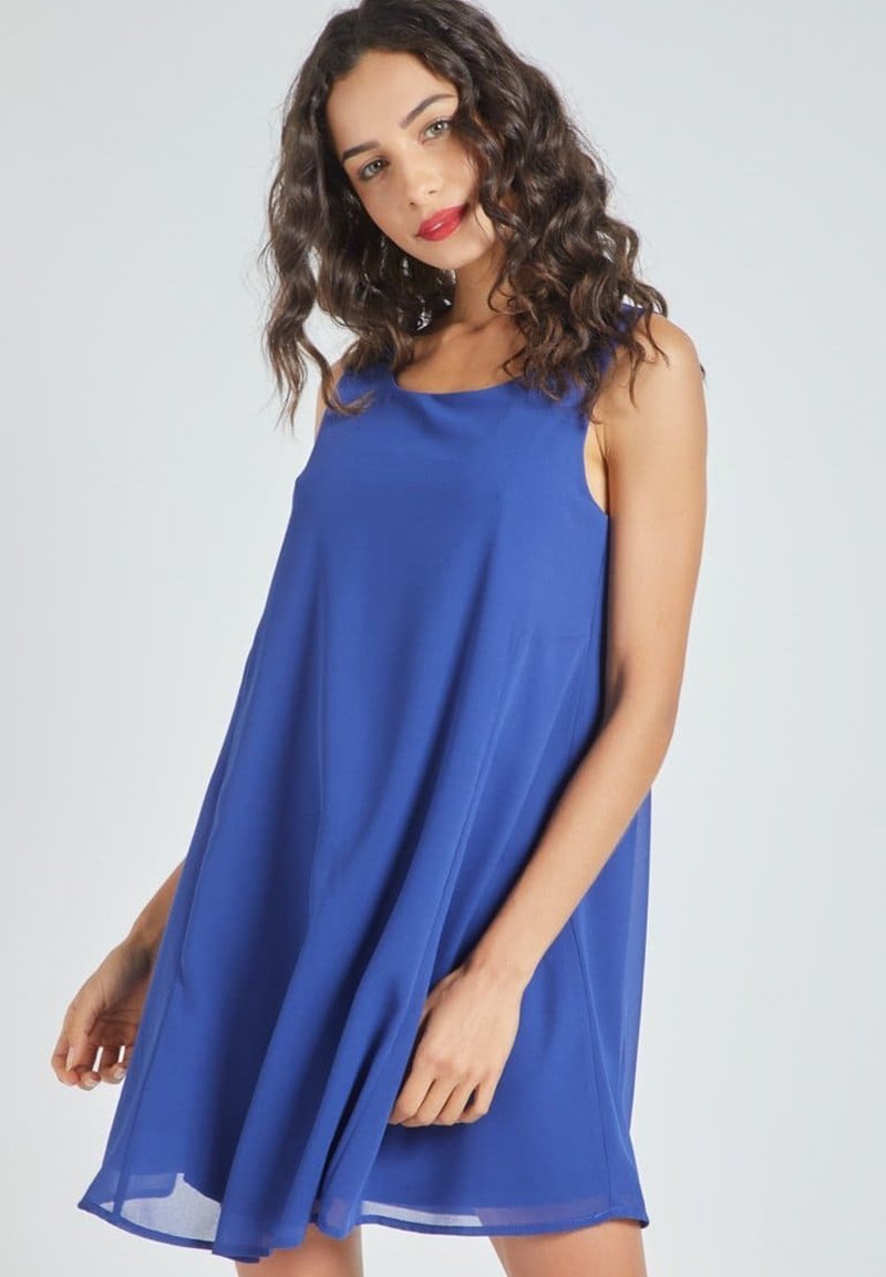 Mujer con cabello rizado, vistiendo un vestido azul sin mangas y de corte suelto, de pie frente a un fondo gris claro.