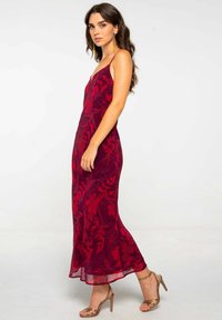Largo vestido rojo ajustado con un patrón floral, hecho de material transparente. Tirantes tipo espagueti y un dobladillo fluido. Combinado con sandalias de tacón metálicas.