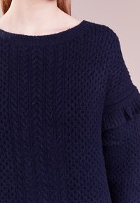 Maglione in blu navy lavorato a maglia con motivo testurizzato, scollo ampio e bordi arricciati sulle maniche. Il materiale sembra morbido e accogliente.