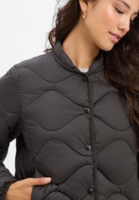 Gepolsterte dunkelgraue Steppjacke mit Stehkragen, schwarzen Druckknöpfen und strukturiertem, wellenförmigem Muster mit glatter Oberfläche.