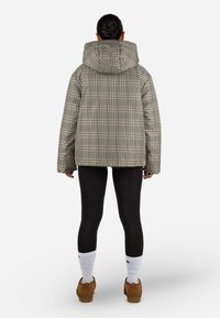 Giacca imbottita a quadri grigia e beige con un grande cappuccio, indossata sopra leggins neri e scarpe marroni, vista da dietro.