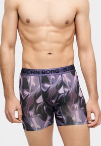 Sportlicher männlicher Oberkörper trägt lila-schwarz gemusterte Boxershorts mit „BJÖRN BORG“-Bund und steht vor einfarbigem Hintergrund.