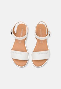 Sandalias blancas de Calvin Klein Jeans con correas en el tobillo y hebilla en tono plateado, suela plana y plantilla beige, vistas desde arriba sobre un fondo blanco.