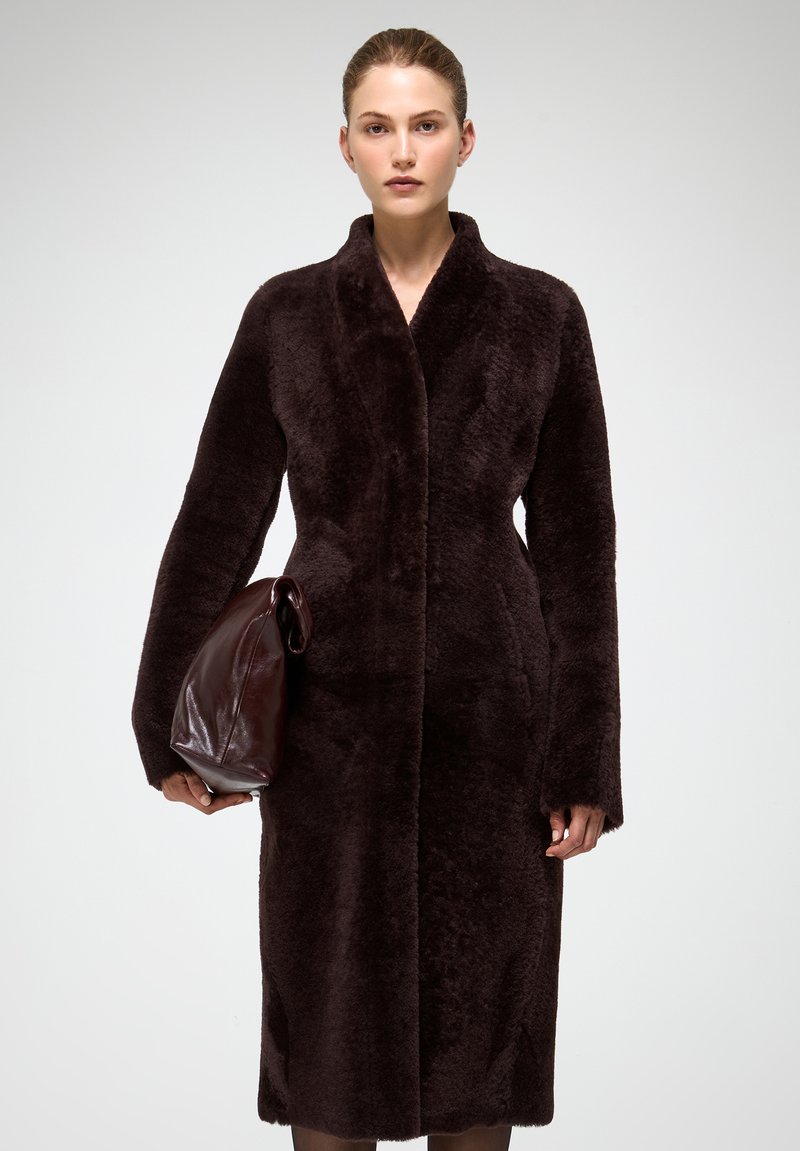 Tumši brūna, mīkstas mākslīgās kažokādas mētelis ar šāla apkakli, garām piedurknēm un taisnu siluetu. Modelis tur tumši brūnu, mīkstu somu.