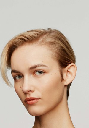 Acercamiento a una persona con cabello rubio claro y corto. Tienen la piel pálida, ojos verdeazulados y llevan un pequeño pendiente de aro dorado.