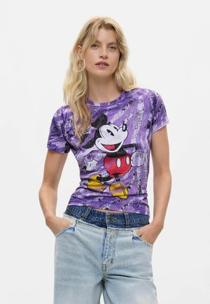 Mujer rubia con camiseta morada de Mickey Mouse y vaqueros azul claro con las manos en los bolsillos, de pie frente a un fondo blanco.