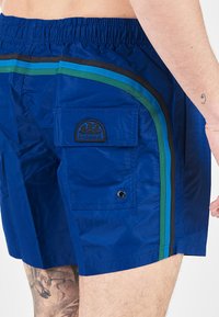 Costumi da bagno blu con vita elastica, tasche laterali e strisce multicolori in verde, blu e nero. Toppa con logo sulla tasca.