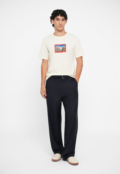 Cremefarvet bomulds t-shirt med et bjerggrafik indrammet i rødt, parret med marineblå stribede bukser. Skoene er lyse med et enkelt design.
