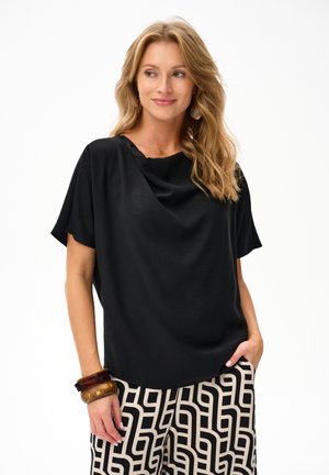 Blondynka w luźnej czarnej bluzce z krótkim rękawem, czarno-białych spodniach w geometryczne wzory oraz drewnianych bransoletkach na lewej ręce.