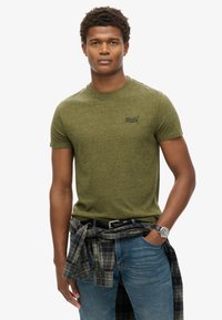 Olivengrøn t-shirt med korte ærmer, rund hals og subtilt logo. Båret sammen med blå jeans og en ternet skjorte bundet omkring livet.