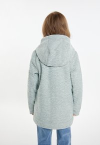 Ljusgrå, texturerad hoodie med stor huva, långa ärmar och en något längre bakre fåll, tillsammans med blå jeans.