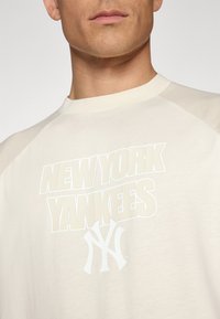 Kremfarget t-skjorte med kort erme og raglanermer, som har stor hvit tekst "NEW YORK YANKEES" og logo på forsiden.