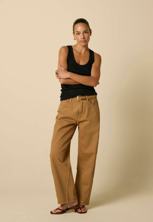Jeune femme debout les bras croisés, portant un haut noir sans manches, un pantalon marron à jambes larges avec ceinture, et des sandales marron à talons.