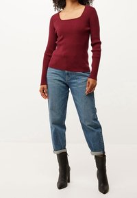 Burgunderfarbener Rippstrickpullover mit quadratischem Ausschnitt, kombiniert mit aufgerollten blauen Jeans und schwarzen Knöchelstiefeletten. Schlichtes, tailliertes Design.