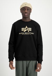 Fekete pulóver arany logóval és "ALPHA INDUSTRIES" felirattal az elején. Kerek nyakkal és bordás mandzsettával rendelkezik, zöld nadrággal viselték.