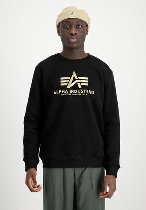 Zwarte sweatshirt met gouden logo en tekst "ALPHA INDUSTRIES" op de voorkant. Heeft een ronde hals en geribde manchetten, gedragen met groene broek.
