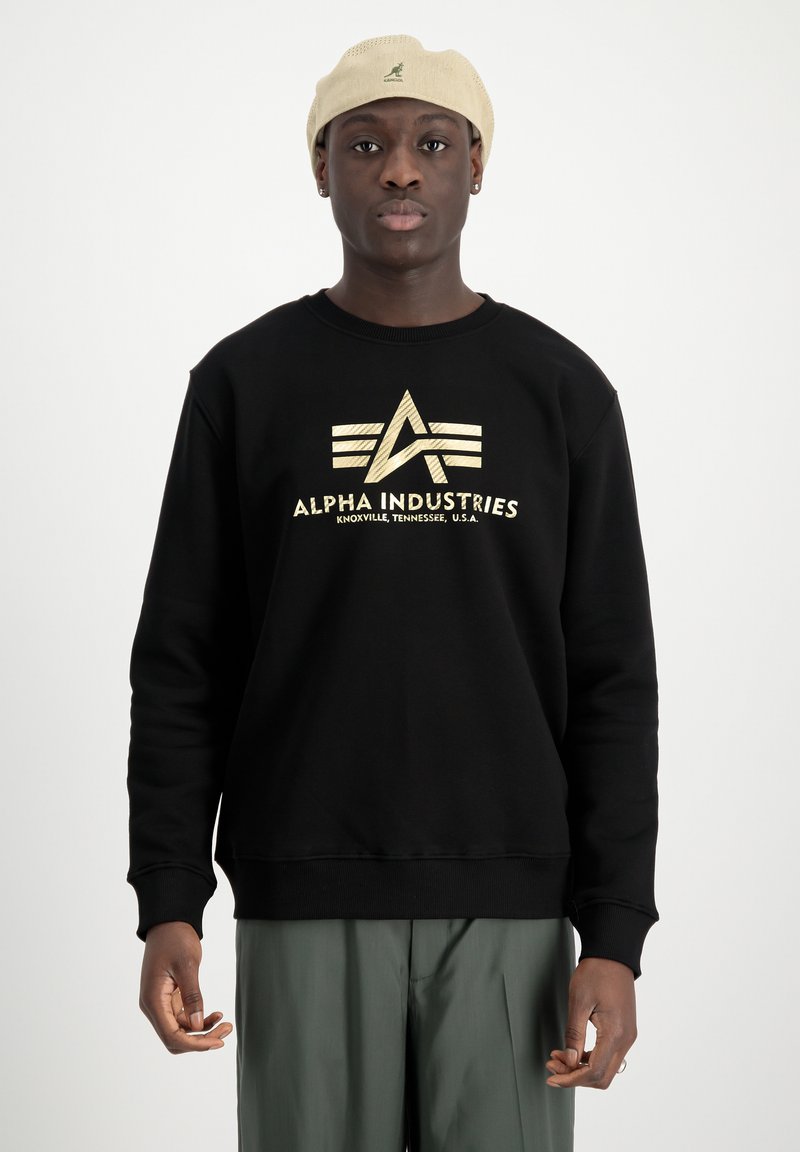 Fekete pulóver arany logóval és "ALPHA INDUSTRIES" felirattal az elején. Kerek nyakkal és bordás mandzsettával rendelkezik, zöld nadrággal viselték.