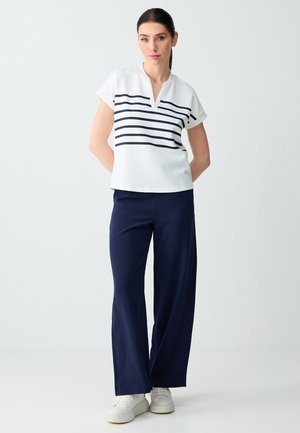 Kobieta ubrana w biały krótki top w granatowe paski, granatowe spodnie o szerokich nogawkach oraz białe trampki, stojąca na jednolitym jasnym tle.