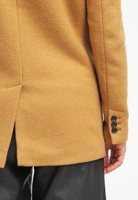 Blazer en laine de chameau avec détail fente au dos, boutons noirs aux poignets, porté avec un pantalon en cuir noir, montrant la vue latérale et arrière du torse et du bras.