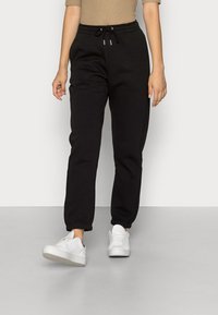 Pantalones jogger de algodón negro con cintura elástica y cordón. Presenta puños acanalados y un ajuste relajado. Combinados con zapatillas blancas.