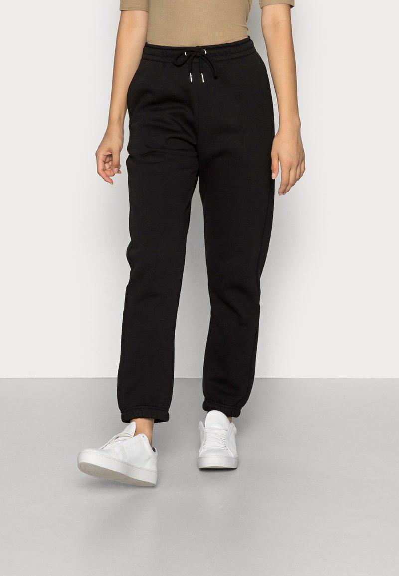 Pantalones jogger de algodón negro con cintura elástica y cordón. Presenta puños acanalados y un ajuste relajado. Combinados con zapatillas blancas.