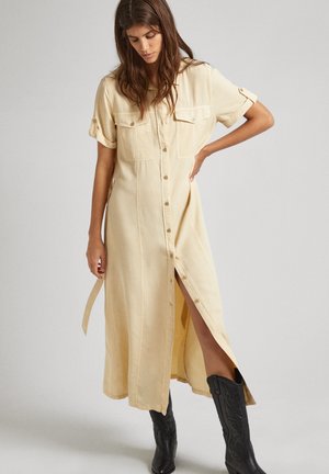Vestido de camisa largo beige hecho de tela ligera, con mangas cortas enrolladas, botonera delantera y aberturas laterales. Combinado con botas de cowboy negras.