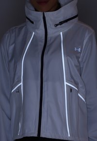 Osoba nosząca jasno szarą kurtkę z kapturem, z pionowymi odblaskowymi paskami oraz logo Under Armour na piersi, zapinaną na zamek.