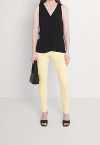 Žena v černém bezramenném topu s V výstřihem, světle žlutých úzkých kalhotách, černých sandálech na podpatku a držící černou tkanou kabelku.