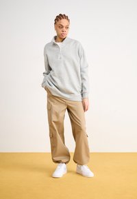 Lys grå hettejakke med glidelås, beige cargobukser med lommer, og hvite joggesko. Modellen står på et gult gulv mot en lys bakgrunn.