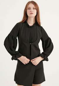 Das schwarze Outfit zeichnet sich durch einen Rüschensaumkragen, texturierte Ärmel, massive Manschetten und einen Reißverschluss vorne aus, mit einem figurbetonten Design und einem knielangen Rock.