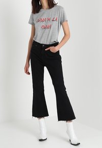 Person iført en grå t-shirt med grafik, sorte cropped flare jeans med frynser og hvide ankelstøvler, stående med den ene hånd i lommen.