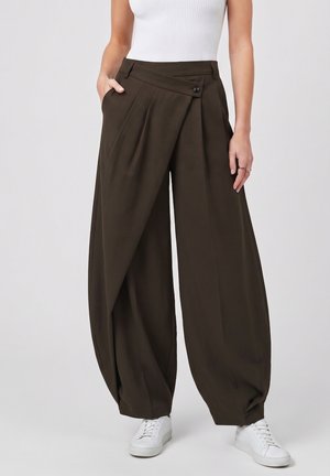 Mujer con pantalones marrón oscuro de talle alto y pierna ancha con solapa frontal asimétrica y zapatillas blancas, de pie con una mano en el bolsillo.