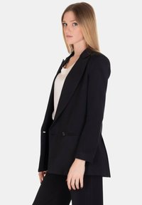 Blazer nero con una vestibilità su misura, revers a punta, chiusura con un bottone e due tasche frontali. Indossato sopra una maglietta bianca.