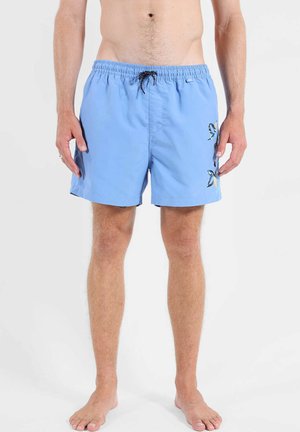 Homme portant un short de bain bleu clair avec un cordon noir et de petites illustrations de poissons sur la jambe droite, debout pieds nus sur fond blanc.
