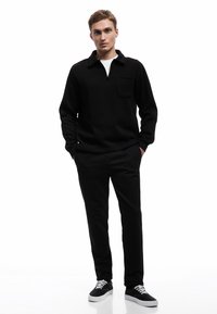 Sudadera negra con media cremallera y bolsillo en el pecho, combinada con pantalones negros y zapatillas negras con cordones blancos. Textura de tela suave.
