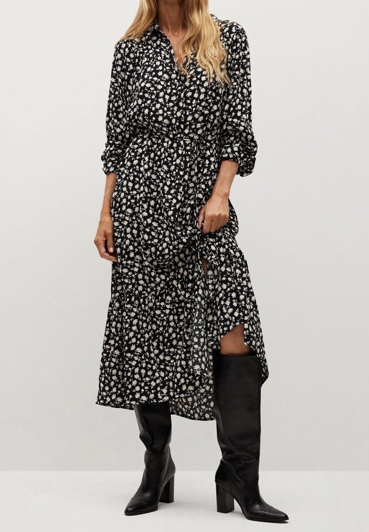 Femme en robe midi noire à fleurs avec taille froncée, associée à des bottes en cuir noires montant jusqu'au genou, debout devant un fond gris uni.