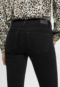 Persona con jeans ajustados negros con bolsillos traseros y una camisa de manga larga con estampado de leopardo, de pie de lado contra un fondo blanco.