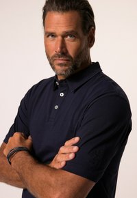 Tummansininen puuvillapolo-paita lyhyillä hihoilla, kolmen napin nappilista ja brodeerattu logo vasemmassa hihassa. Sujuva pinta, klassinen malli.