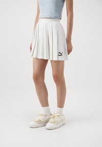 Witte geplooide rok met een hoge taille en een zwart Puma-logo op de zoom, gecombineerd met witte sneakers met beige accenten en dikke veters.